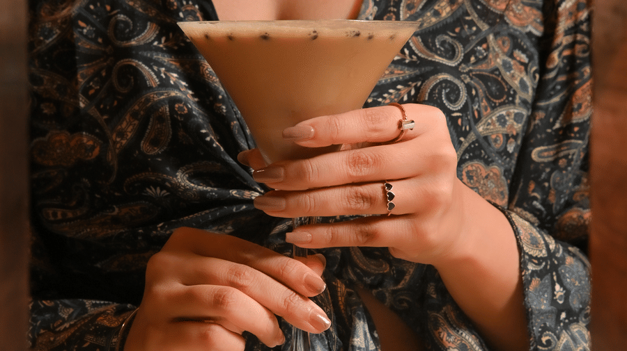 A woman wearing a patterned dress is holding a cocktail glass filled with a creamy beverage, viewed through a wooden-framed mirror - Ananta Spa and Resort, Pushkar.