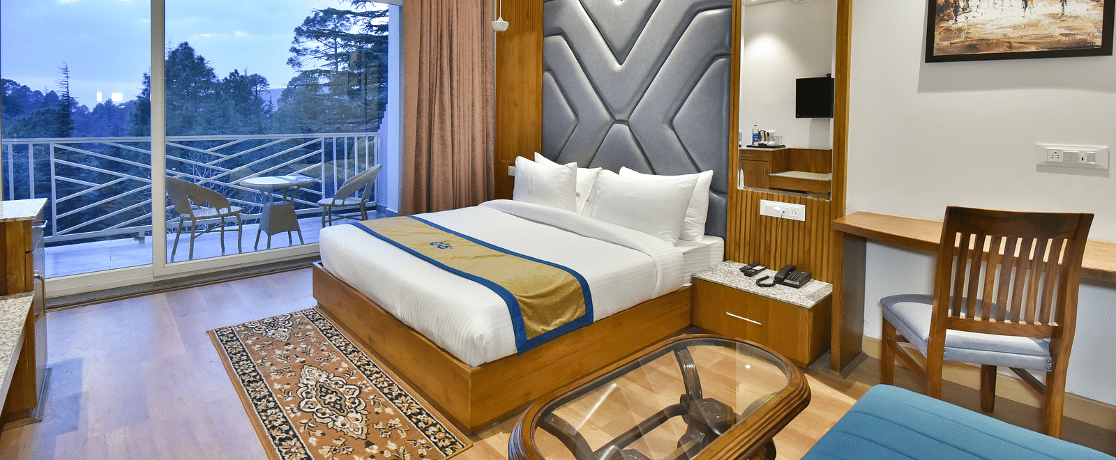 Image of the luxury room with balcony with a bed, bed side tables, chair, work desk, and a glass door leading to the balcony with a scenic view in the background - DLS Ark Holiday Inn, Dalhousie