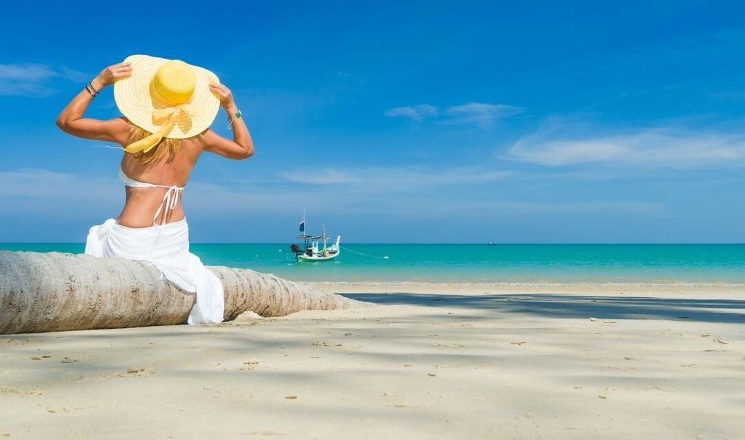 A woman sitting on a fallen trunk of a coconut tree in the sea shore, facing the expansive sea and a yacht.