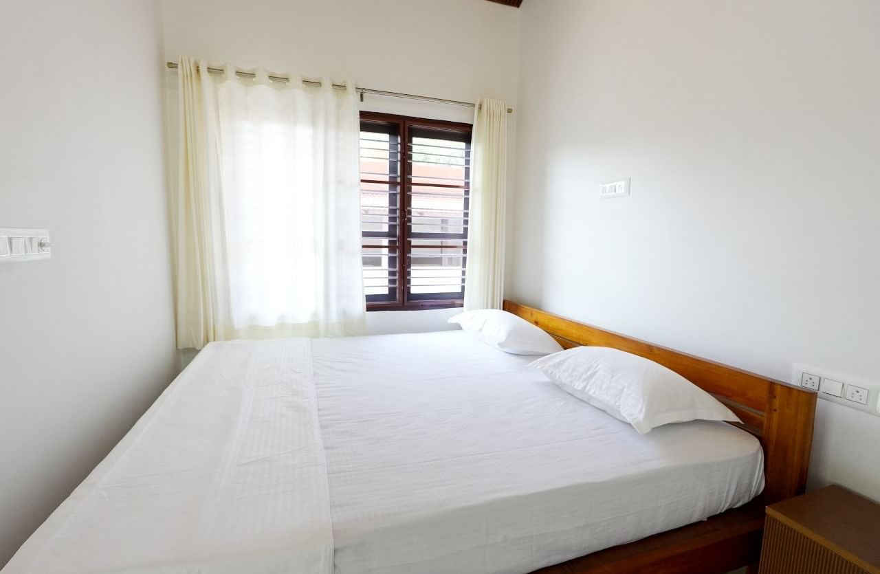 A bright and inviting deluxe room at Bhuthanakadu Retreat, showcasing a comfortable bed with white bedding, a wooden frame, and large windows draped with sheer white curtains.