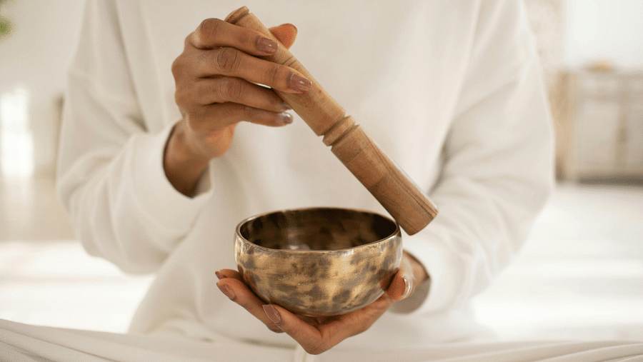Hands in white clothing holding a Tibetan singing bowl and wooden mallet, preparing for a calming sound therapy session.