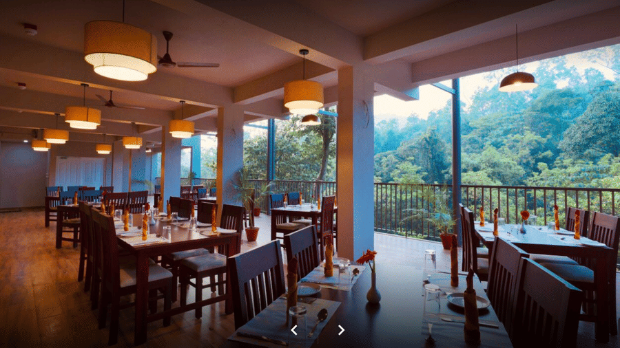 A spacious restaurant with wooden furniture, large windows, and a scenic view of lush greenery outside.- Abad Brookside Lakkidi, Wayanad