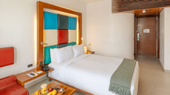 View of a room at The Promenade Pondicherry with a king bed, multicoloured headboard panel, wooden nightstands, fruit tray, and warm ambient lighting.