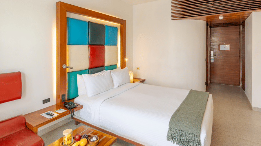 View of a room at The Promenade Pondicherry with a king bed, multicoloured headboard panel, wooden nightstands, fruit tray, and warm ambient lighting.