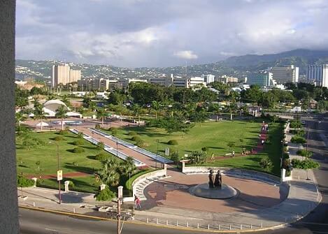 An aerial view shows a lush green park area with roads and buildings in the background under a cloudy sky.