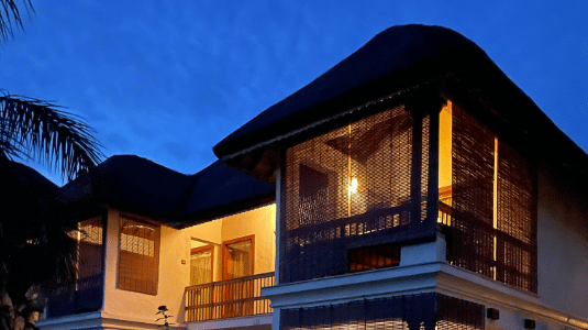 An exterior view of a 2-storey resort villa illuminated at night, featuring wooden balconies and a lawn | Le Pondy