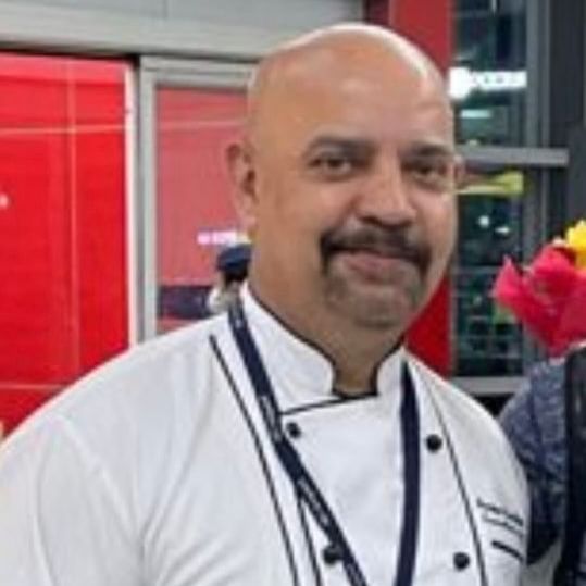 Portrait of Sanjay Gambhir in a white chef coat with a lanyard, standing in front of a red background.