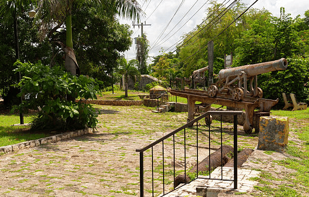An outdoor view of the Trench Town Culture Yard Museum in Kingston, Jamaica, featuring a rustic yard space with preserved structures, overhead shade canopies, and lush surrounding tropical vegetation evoking the community's deep-rooted heritage.