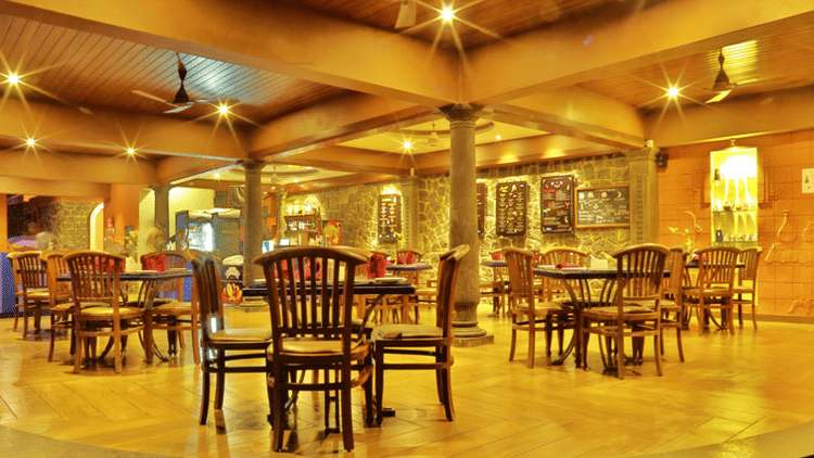 The warmly lit Lounge of Uday Samudra Leisure Beach Hotel features wooden tables and chairs arranged on a raised platform with pillars.