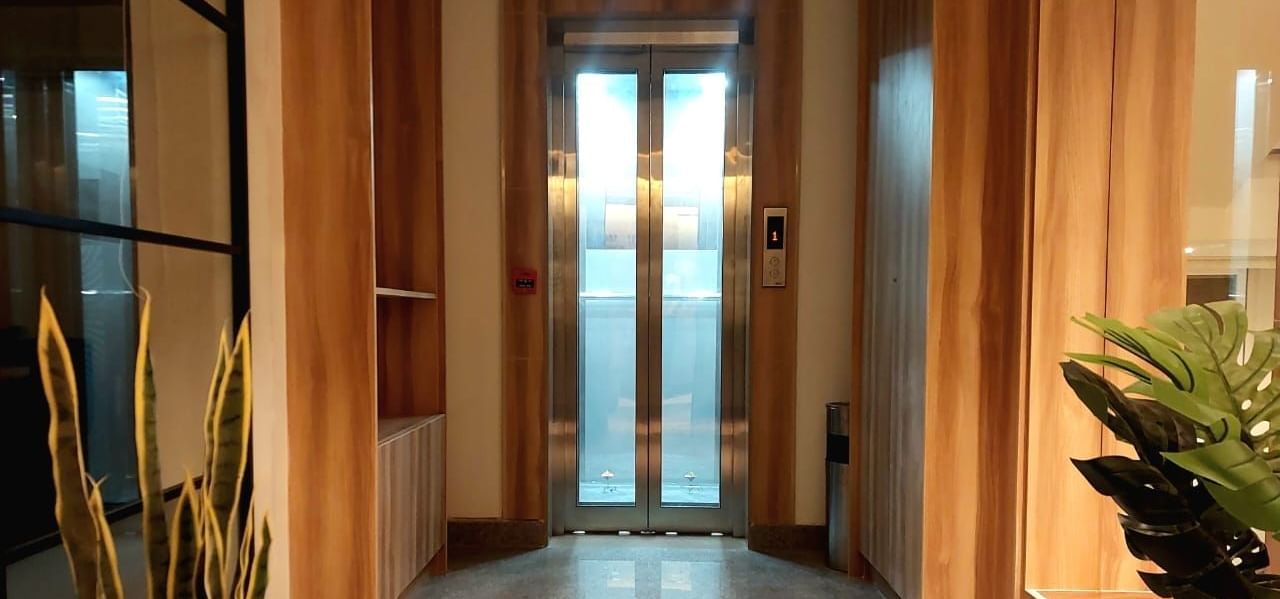 An elevator with its door closed at the end of the corridor at Hotel Raai, Hosur. The corridor has a few flower pots like snake plant and monstera plant.