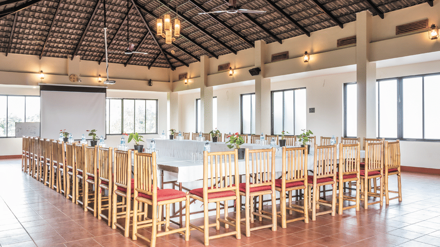A long, elegantly set table in the event hall at Forest Escapes Koyna, with a ceiling fans, chandelier, projector, and a screen in view. Forest Escapes is the best place for a corporate team outing near Pune.