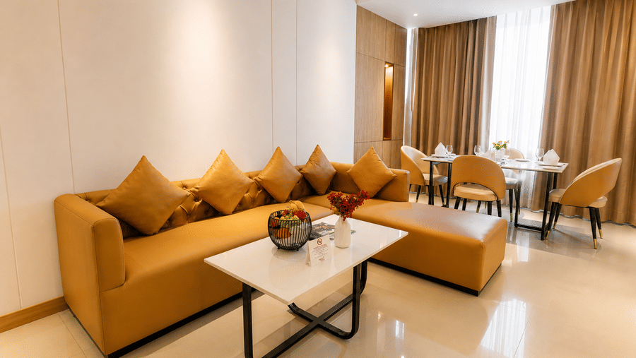 A living and dining area inside a suite at Pride Premier Greater Noida, featuring an L-shaped sofa, a coffee table, and a dining table with chairs.