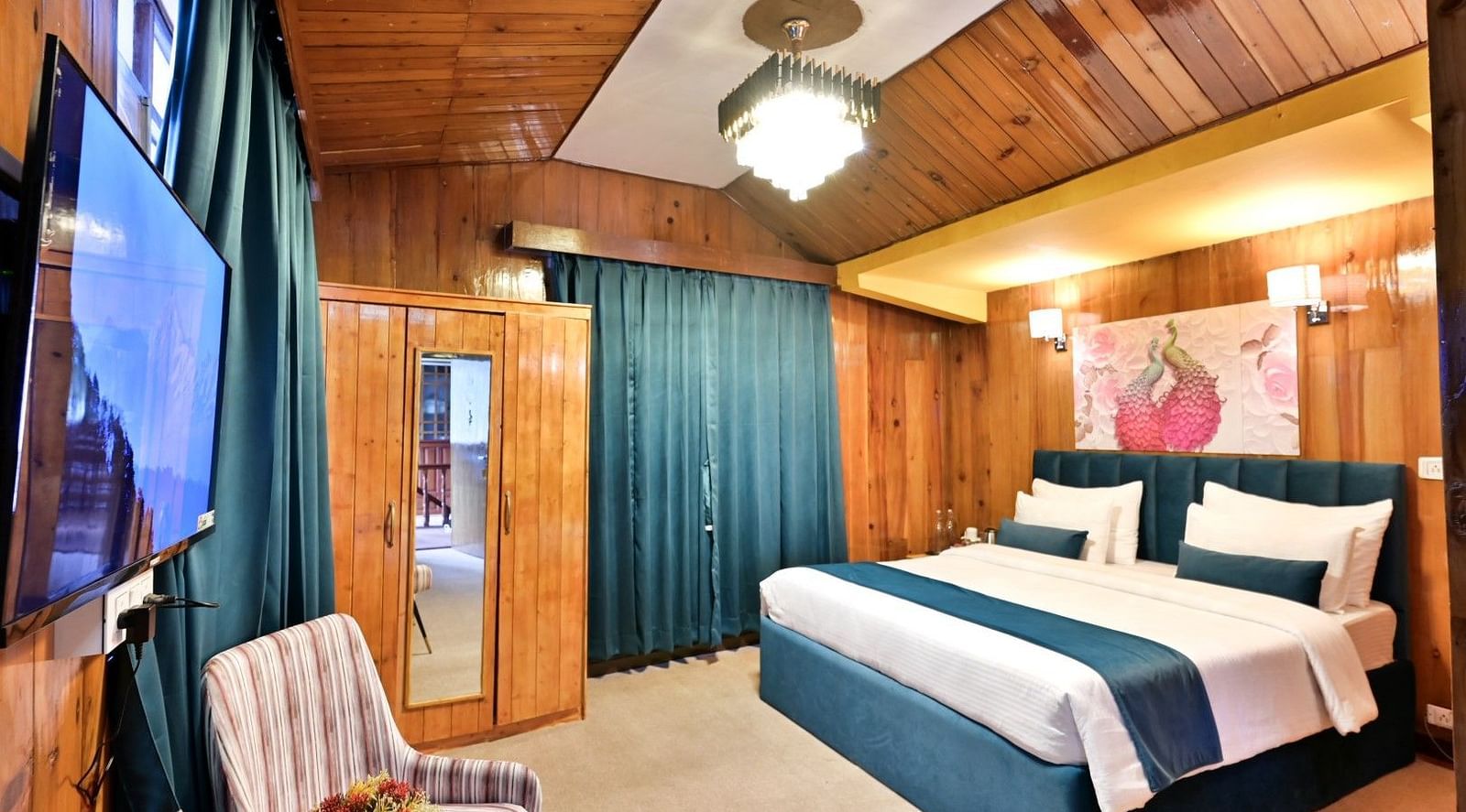 A cosy and well-furnished hotel room with a comfortable bed, a wardrobe, a television, and a seating area with a chair and a table.