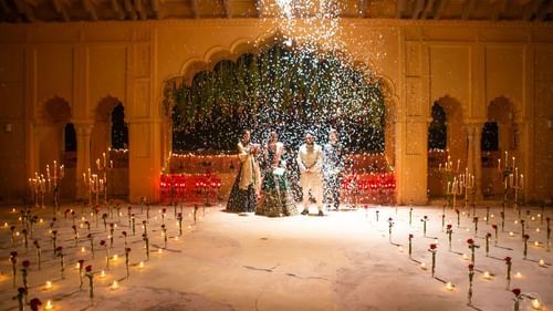 Four people stand under an archway with a shower of particles falling on them, in a space marked with candles and rose stems, at Tijara Fort-Palace - 19th Century, Alwar.