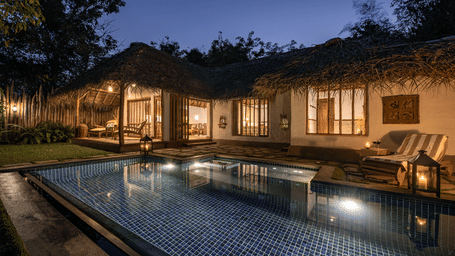 A luxury Earthitects villa with a thatched roof and a private swimming pool glowing under the evening sky.