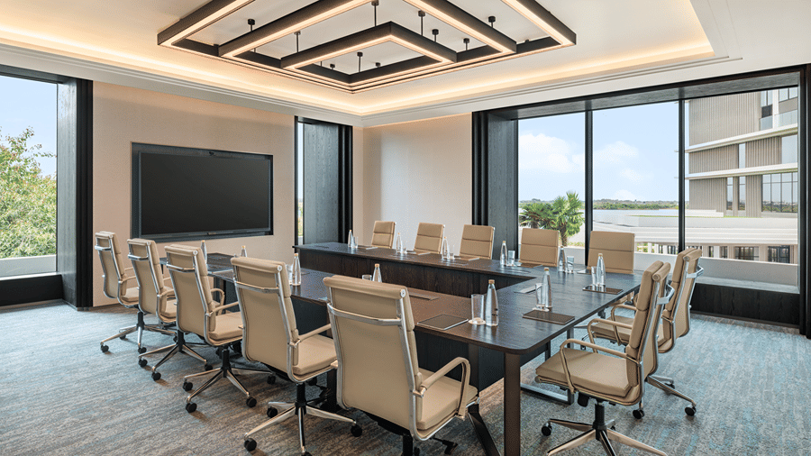 Origin 1 boardroom with a long table, beige leather executive chairs, a wall-mounted screen and floor-to-ceiling windows at The Residency Signature Madurai.