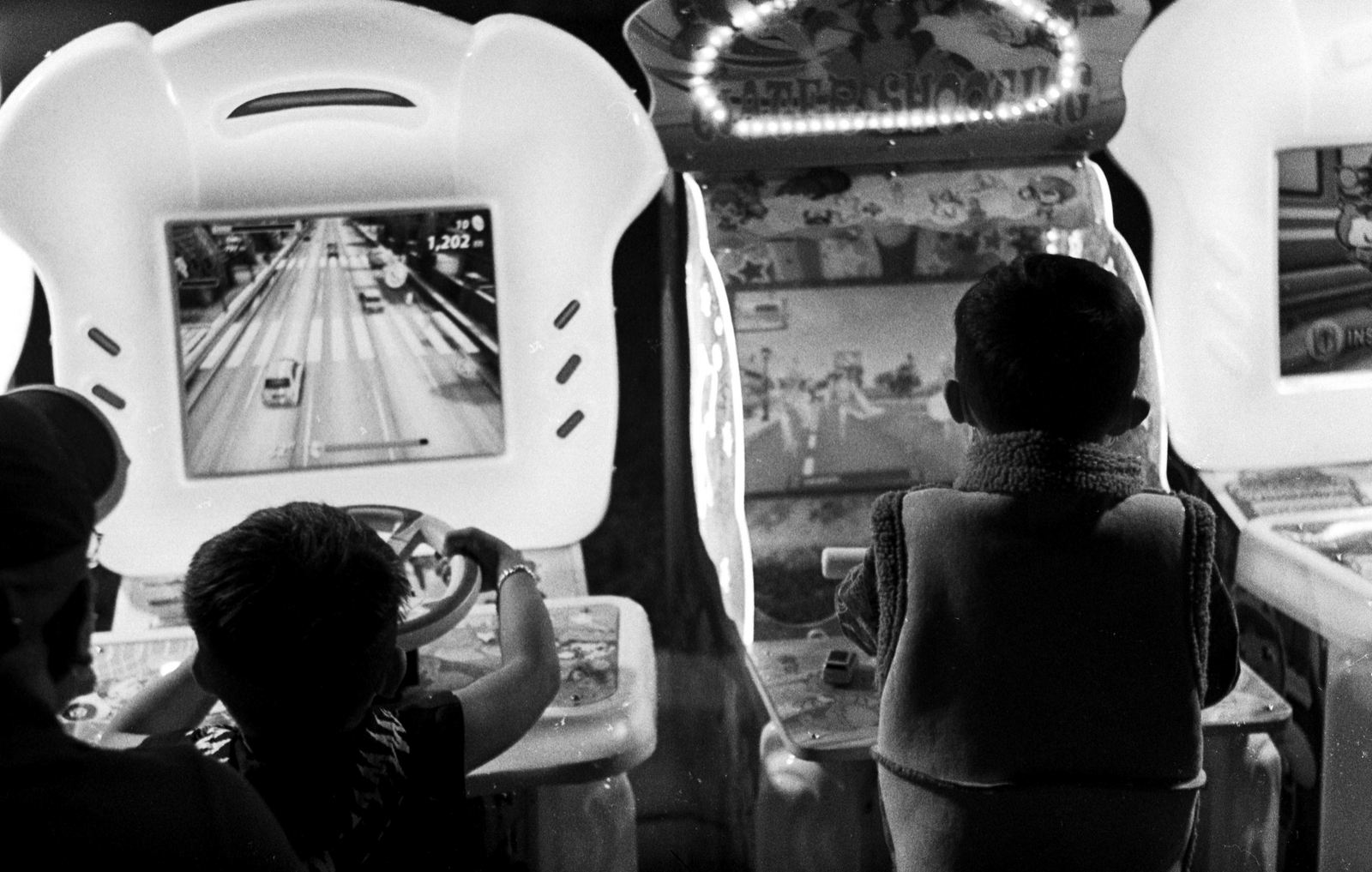 Children playing on arcade racing game machines with steering wheels and screens showing road scenes in a gaming zone.