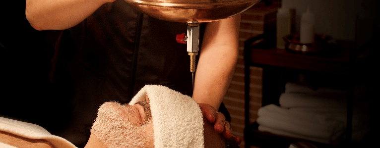 A close-up shows a man lying down with a towel on his forehead, receiving a shirodhara treatment with warm oil pouring from a brass pot at YO1 Longevity & Health Resorts