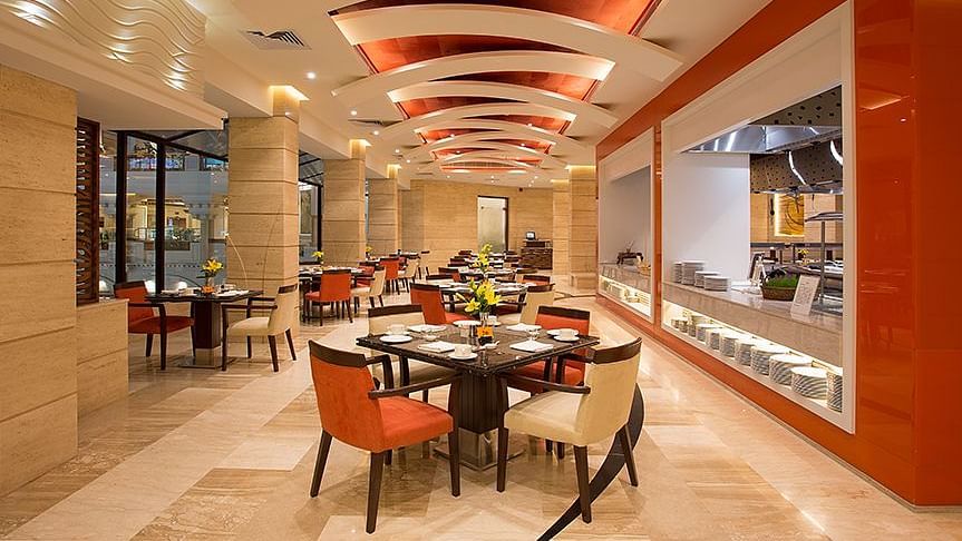 An image of a well lit dining hall where several sets of dining table and chairs are placed - The Suryaa New Delhi