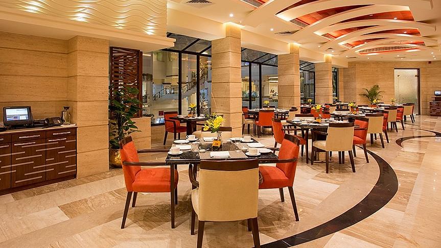 An image of a well lit dining hall where several sets of dining table and chairs are placed - The Suryaa New Delhi