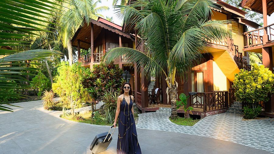 Woman in a blue dress standing in a resort with a suitcase, palm trees in the background under a clear sky.