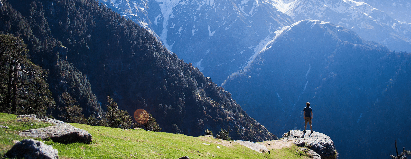 An image of snow caped mountains where a person is seen standing near a cliff enjoying the view