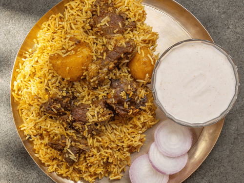 Kolkata style potato biryani served on a plate with raita and sliced onions.