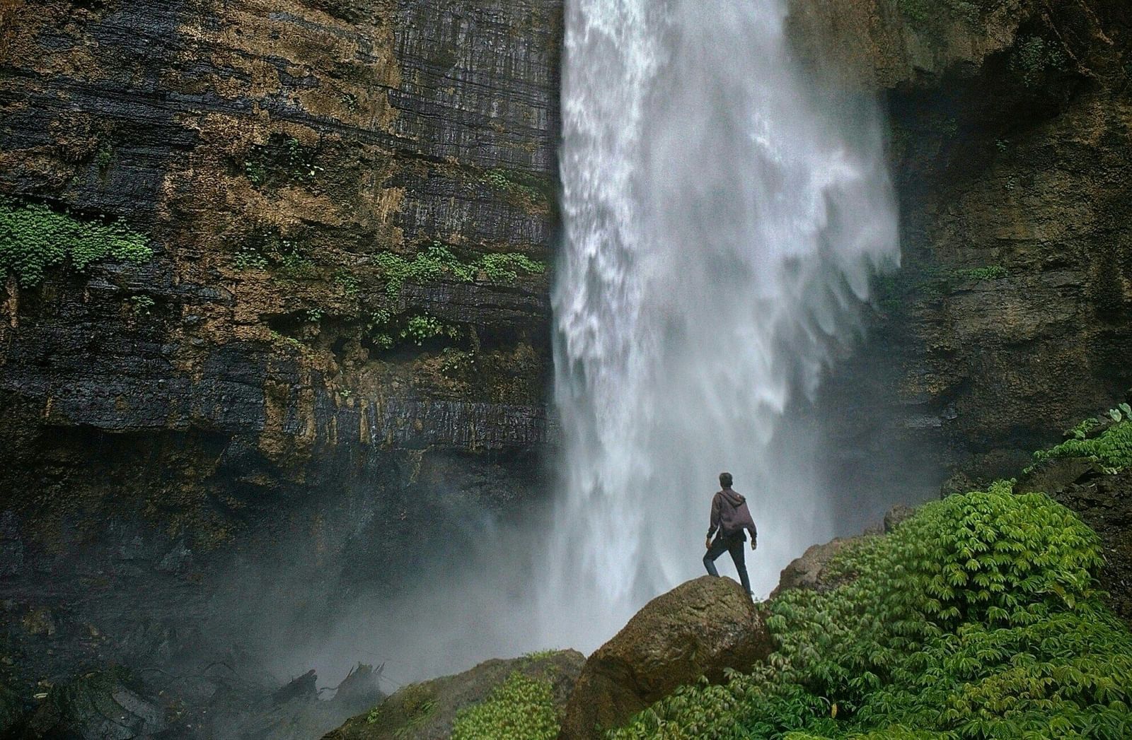 A stunning, close-up, low-angle shot of a powerful waterfall cascading down a rocky cliff face, with a person standing at the bottom for scale.