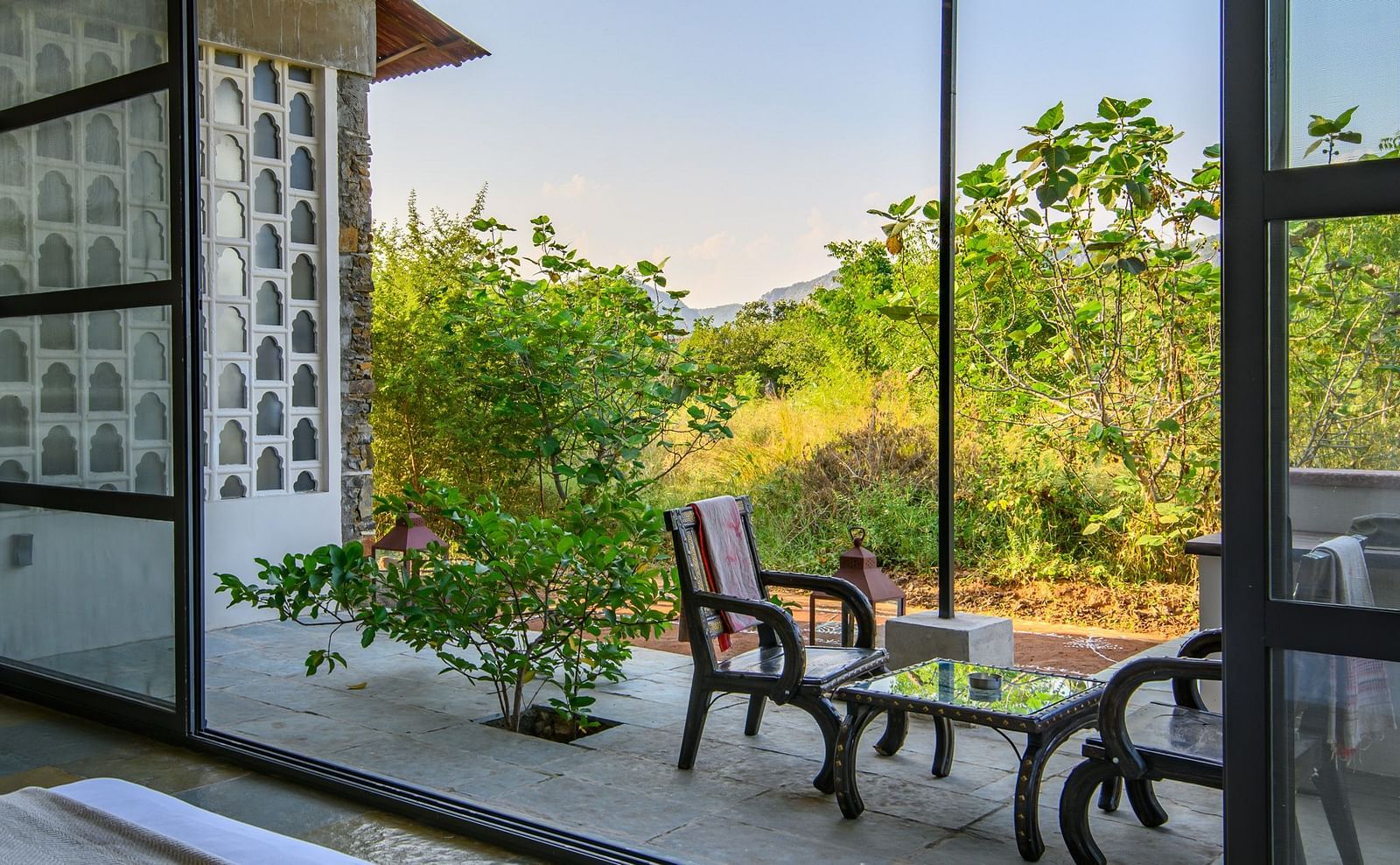An image of a room with a queen size bed, chandelier, ceiling fan, other decorative items and an expansive glass door leading out - Utsav Camp Sariska