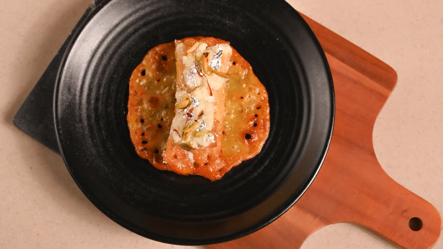 Top-down view of a plated dish with sauce and garnish, served on a black plate placed on a wooden serving board, with a white plate and cutlery in the background - Ananta Spa and Resort, Pushkar.