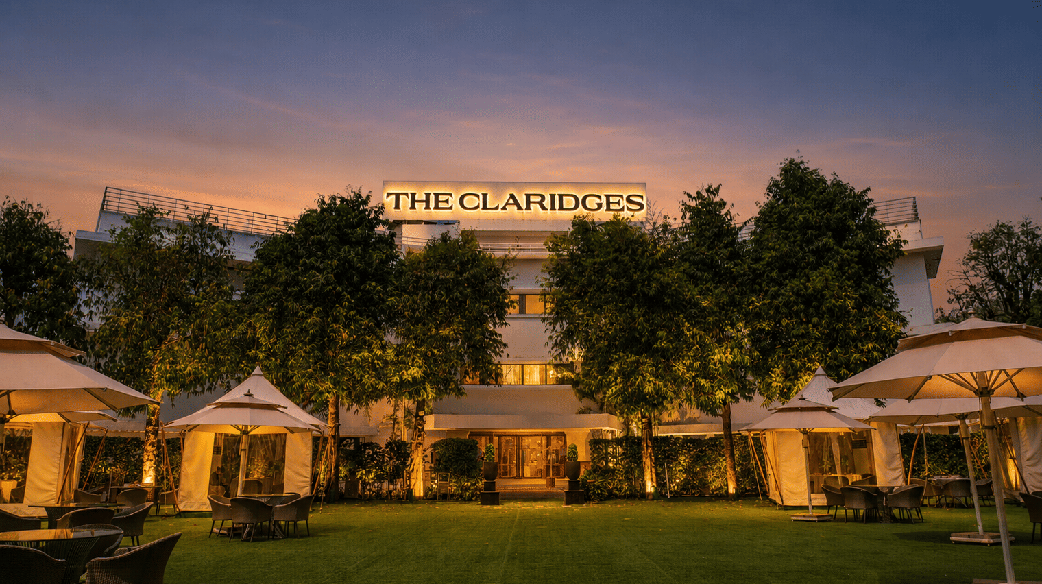 Evening view of a green hotel lawn featuring warm-lit white canopies and outdoor dining tables in front of an illuminated building at dusk - The Claridges New Delhi