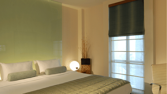 Minimalist bedroom with green quilted bedspread, white pillows with green cushions, green accent wall, window with sheer curtains and roman shade, bedside lamp, and decorative dried plants.