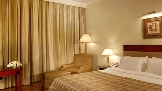 Neatly arranged hotel room with a large bed featuring a checkered bedspread, bedside lamps, cushioned armchair with floor lamp, framed artwork above the headboard, beige curtains, and a small table with a flower arrangement, creating a warm and inviting at