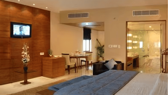 Modern hotel room with a large bed featuring a blue throw blanket, mounted flat-screen TV on a wooden accent wall, decorative vase with flowers, sitting area with table and chairs, and glass-walled bathroom in the background.