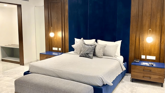 Modern bedroom with a blue bed frame, white and grey bedding, cushioned bench, deep blue wall panel flanked by wooden panels with mounted lights, and white marble flooring.