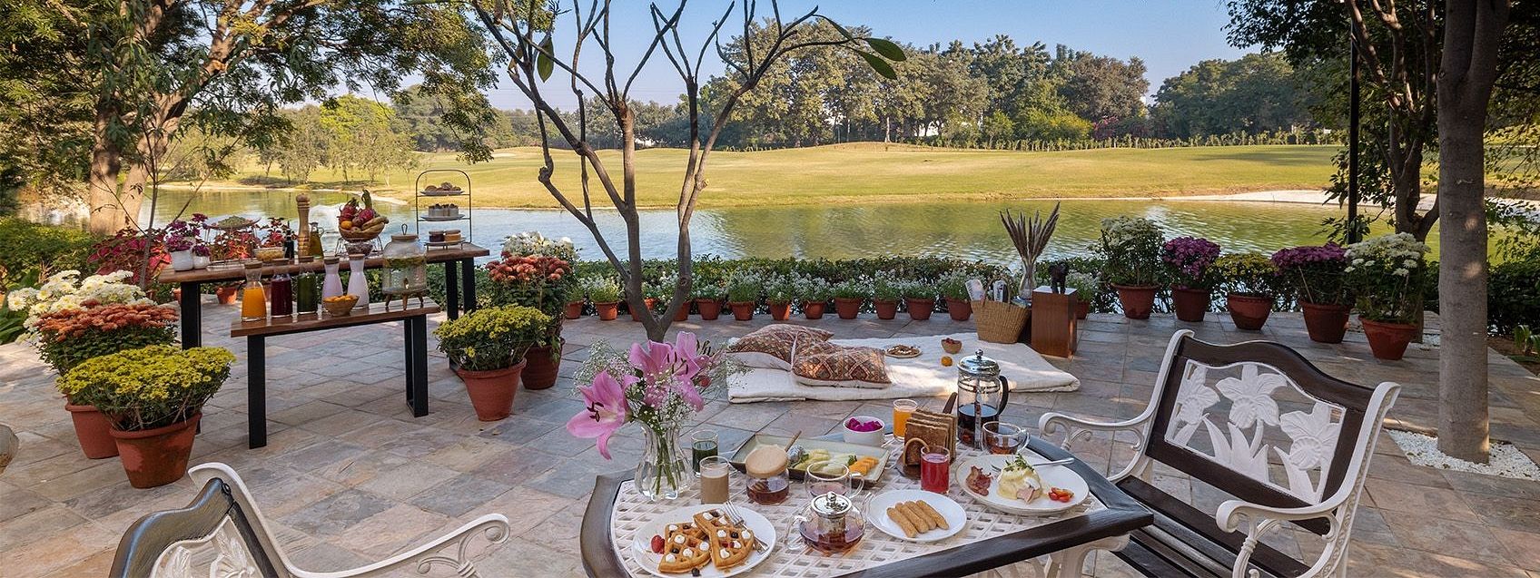 Outdoor dining setup with multiple tables and food spread under trees, overlooking an open green field at Karma Chalets.