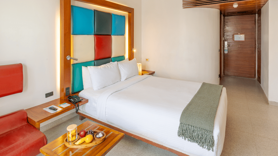 View of a room at The Promenade Pondicherry with a king bed, multicoloured headboard panel, wooden nightstands, fruit tray, and warm ambient lighting.