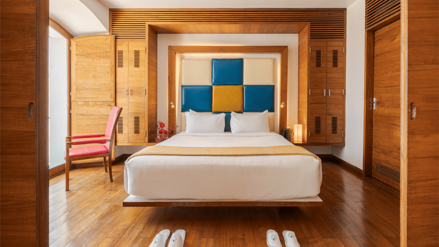 Front view of a suite bedroom at The Promenade Pondicherry with a king bed, blue and yellow headboard, wooden flooring, wardrobe, and soft lighting.