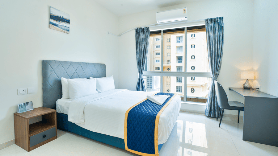A light-filled hotel room with a double bed, blue decorative runner, a simple wooden desk, and a large window with blue curtains at Apartel by Aarin, Oragadam.