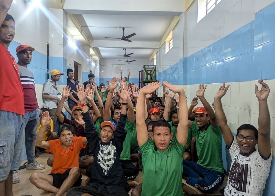 A group of children and young adults with special needs sit indoors, smiling and raising their hands enthusiastically during a group session.