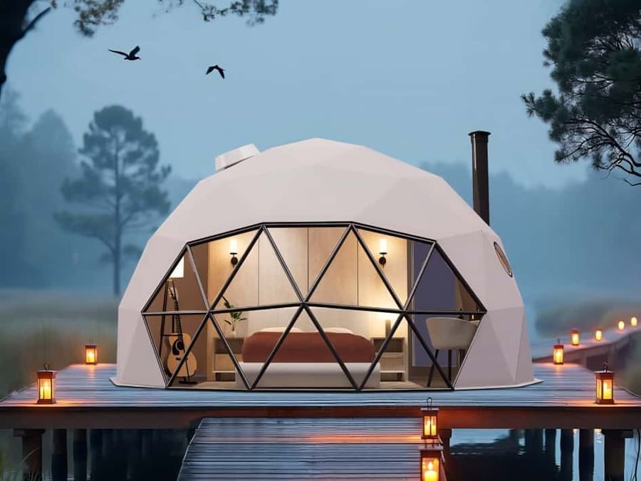 Exterior view of a Glass Dome room at dusk, with ambient lighting and a wooden deck surrounded by trees.