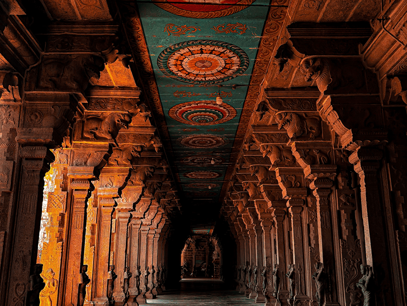 Grand dim corridor inside an Indian temple featuring carved dark pillars and colourful ceiling motifs.