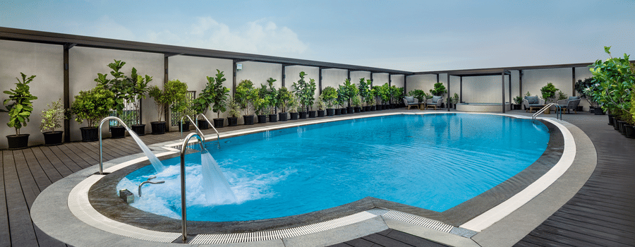 An outdoor swimming pool at The Residency Towers Chennai surrounded by a timber deck and potted plants, with sun loungers visible
