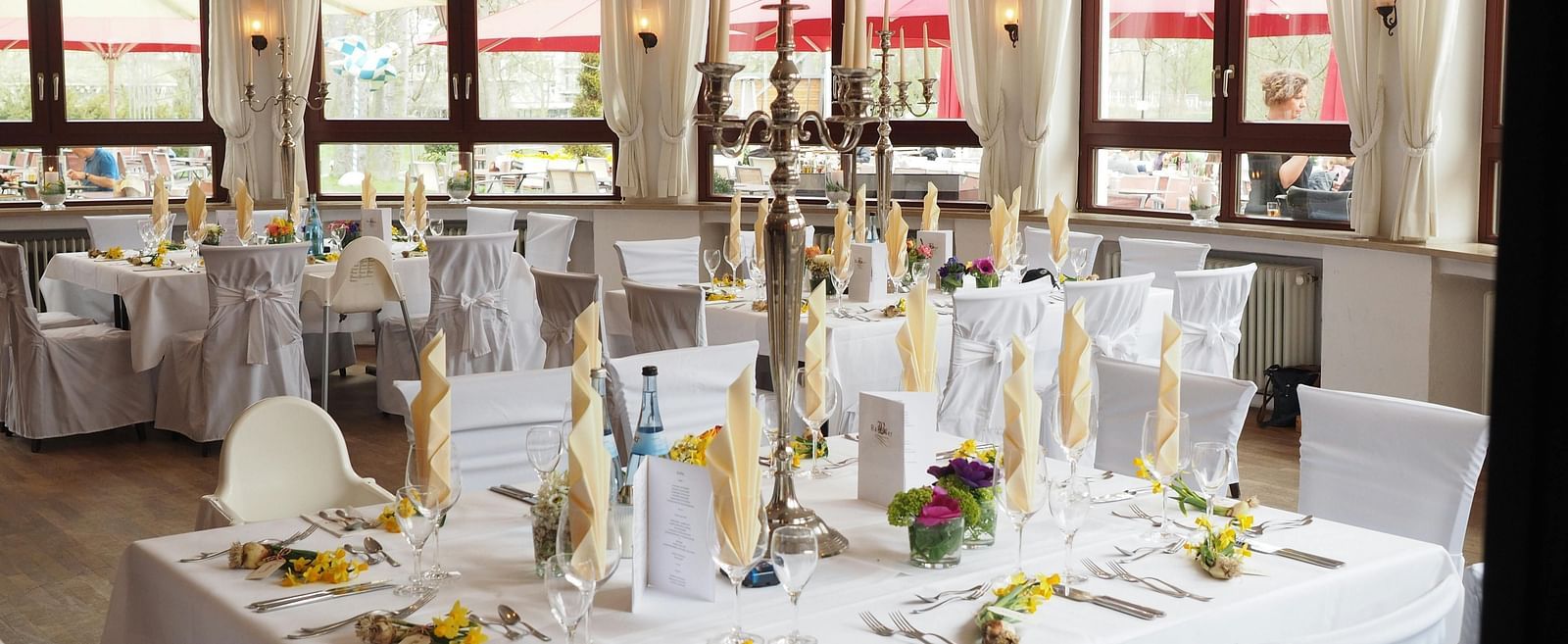 A banquet hall with tables covered with white linens and adorned with floral arrangements, candles, and elegant place settings.