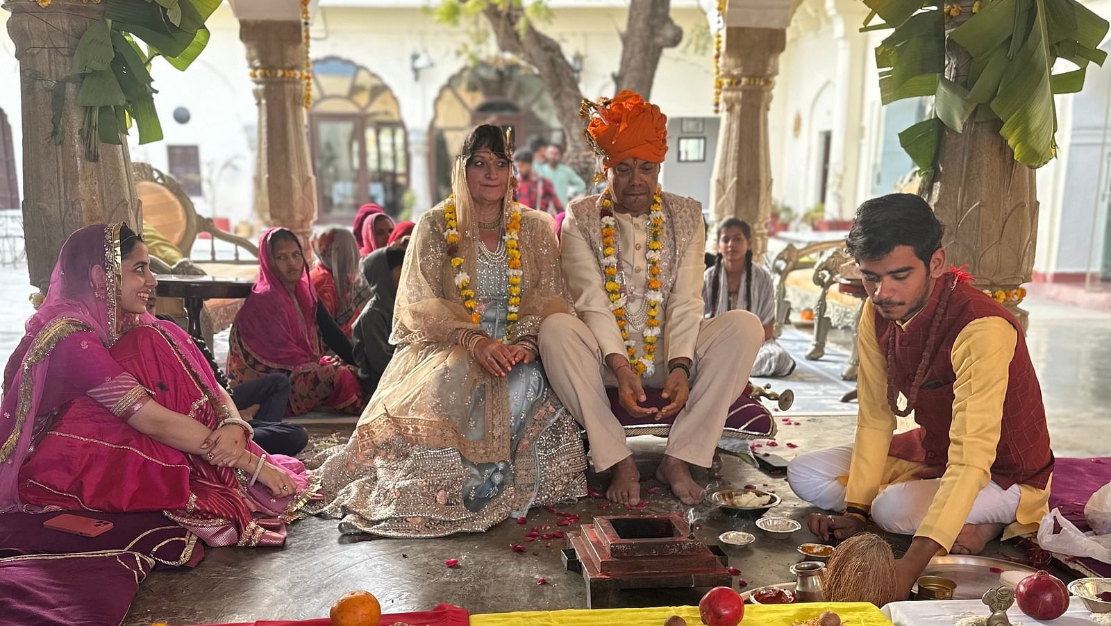 A couple in wedding attire sits at a decorated mandap, surrounded by people and ceremonial items - Fort Barli, Ajmer.