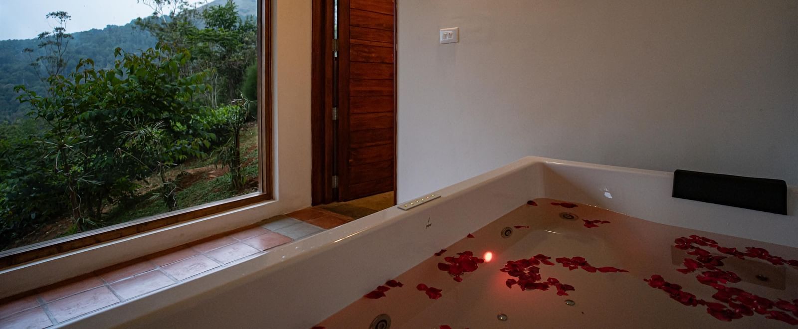 A luxurious bathroom at Vaasta Vagamon with a large tub filled with water and flower petals, beside a window overlooking a natural landscape.