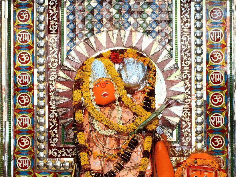 A Hindu deity idol decorated with flower garlands and ornaments, displayed within a colourful shrine framed by detailed geometric patterns.