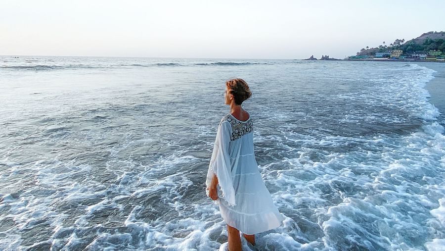 A person in white clothing walks through shallow ocean water, with waves breaking around their legs. The horizon and sky are visible in the background.