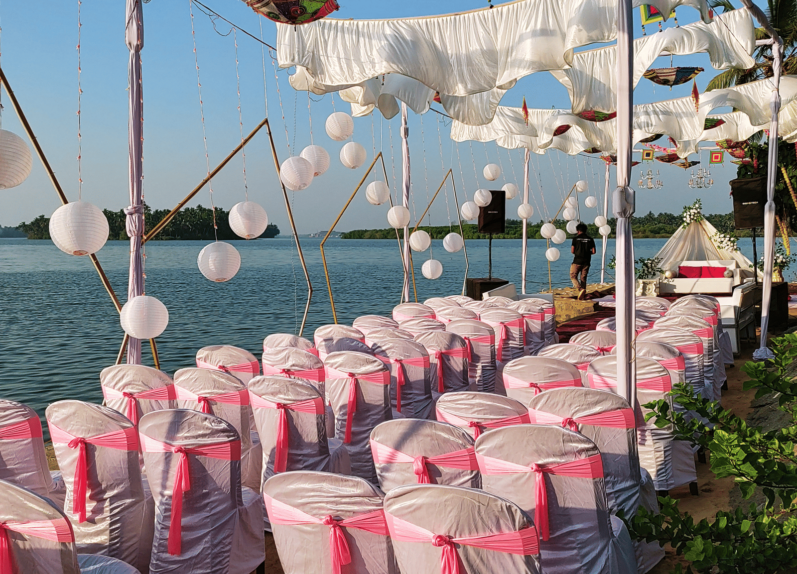 Rows of white chairs with pink covers arranged outdoors under hanging umbrellas at Paradise Lagoon Resort, Udupi.