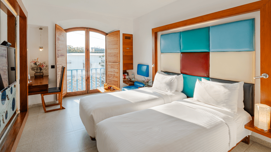 Wide view of a room at The Promenade Pondicherry with twin beds, a multicoloured headboard panel, arched windows, and bright natural light.
